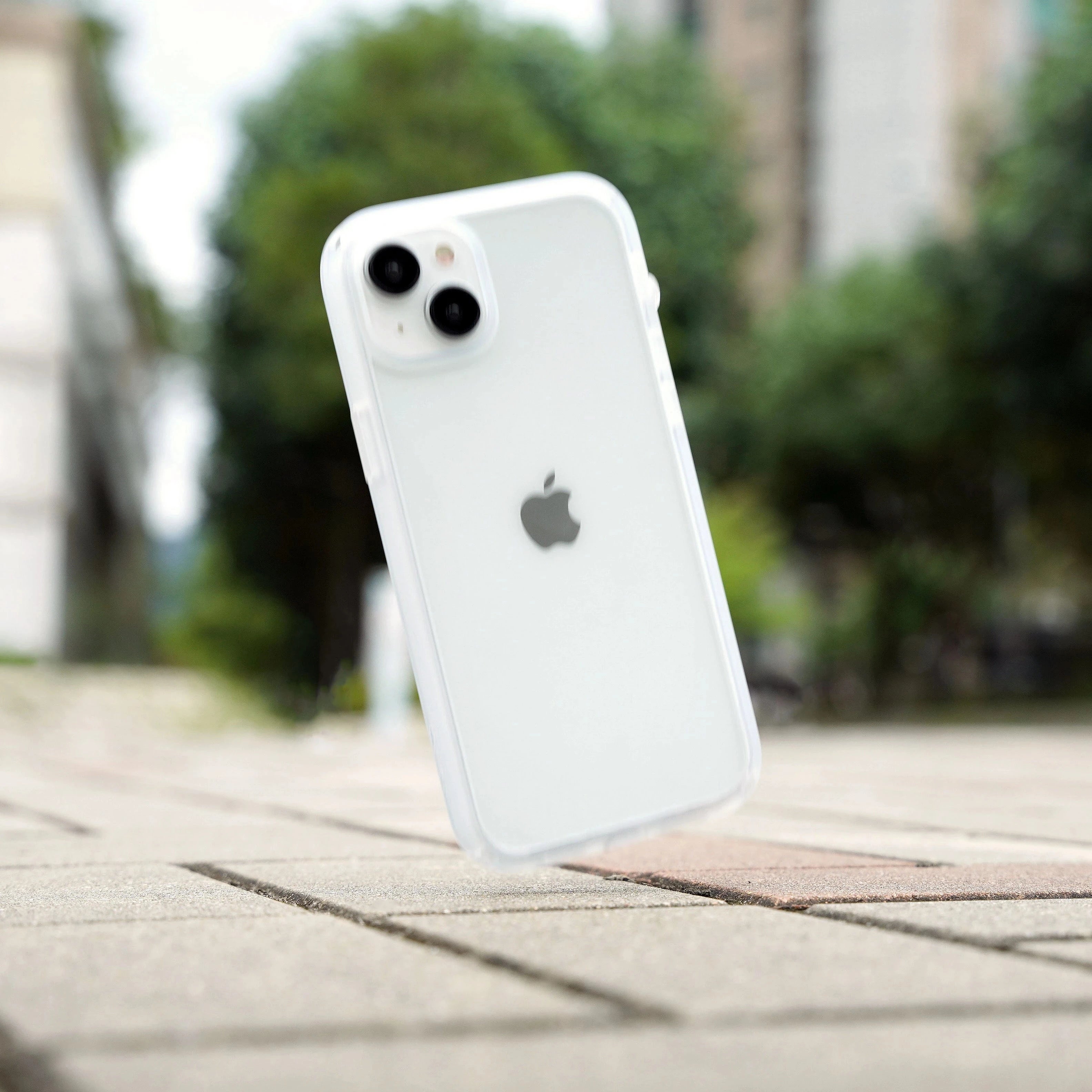 The Catalyst iPhone - Drop Proof Case in clear color makes a white iPhone appear to float above a tiled pavement, with blurred trees and buildings in the background, showcasing its sleek design and robust impact protection.