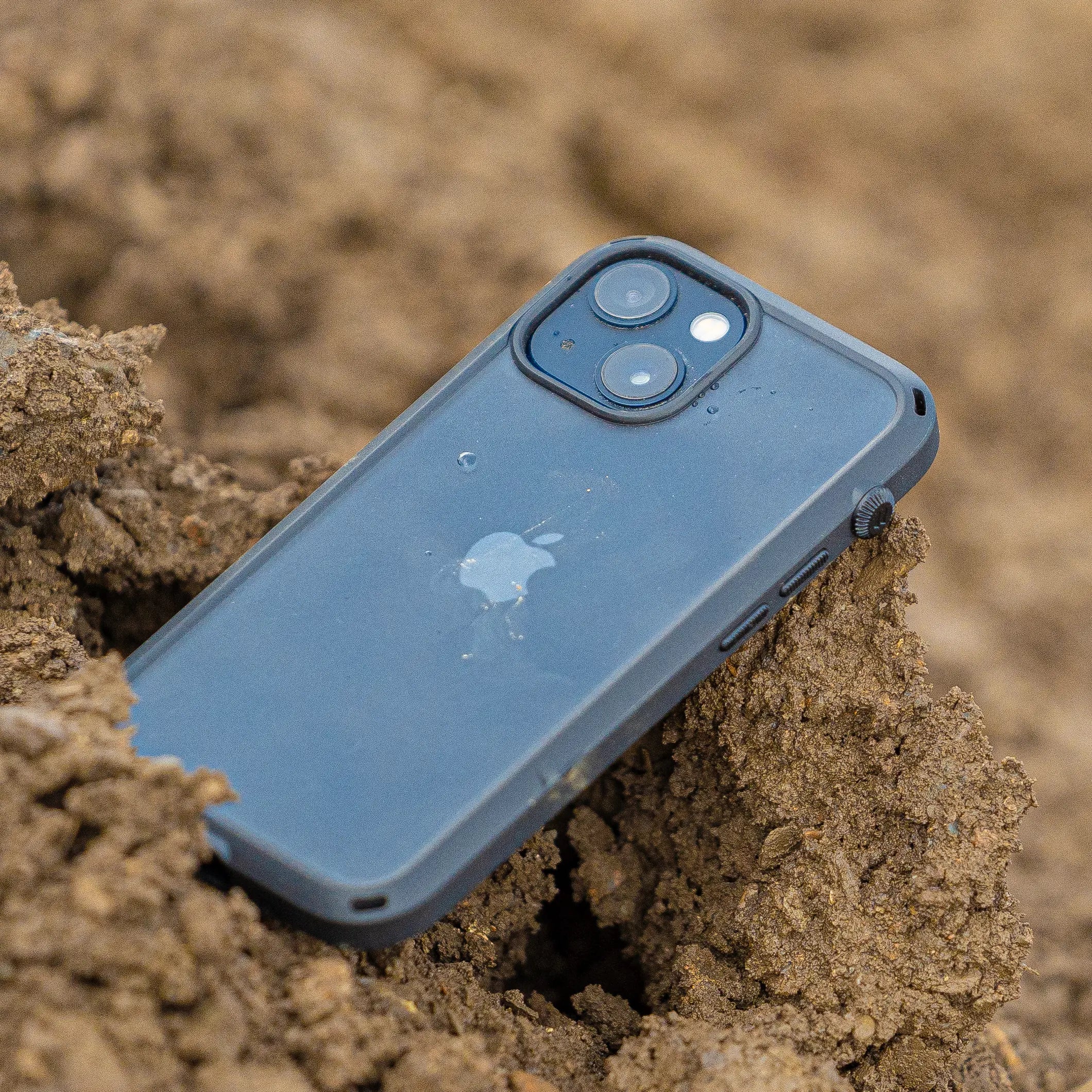 An iPhone with a Catalyst Drop Proof Case in black, featuring dual cameras, is partially buried in soil.