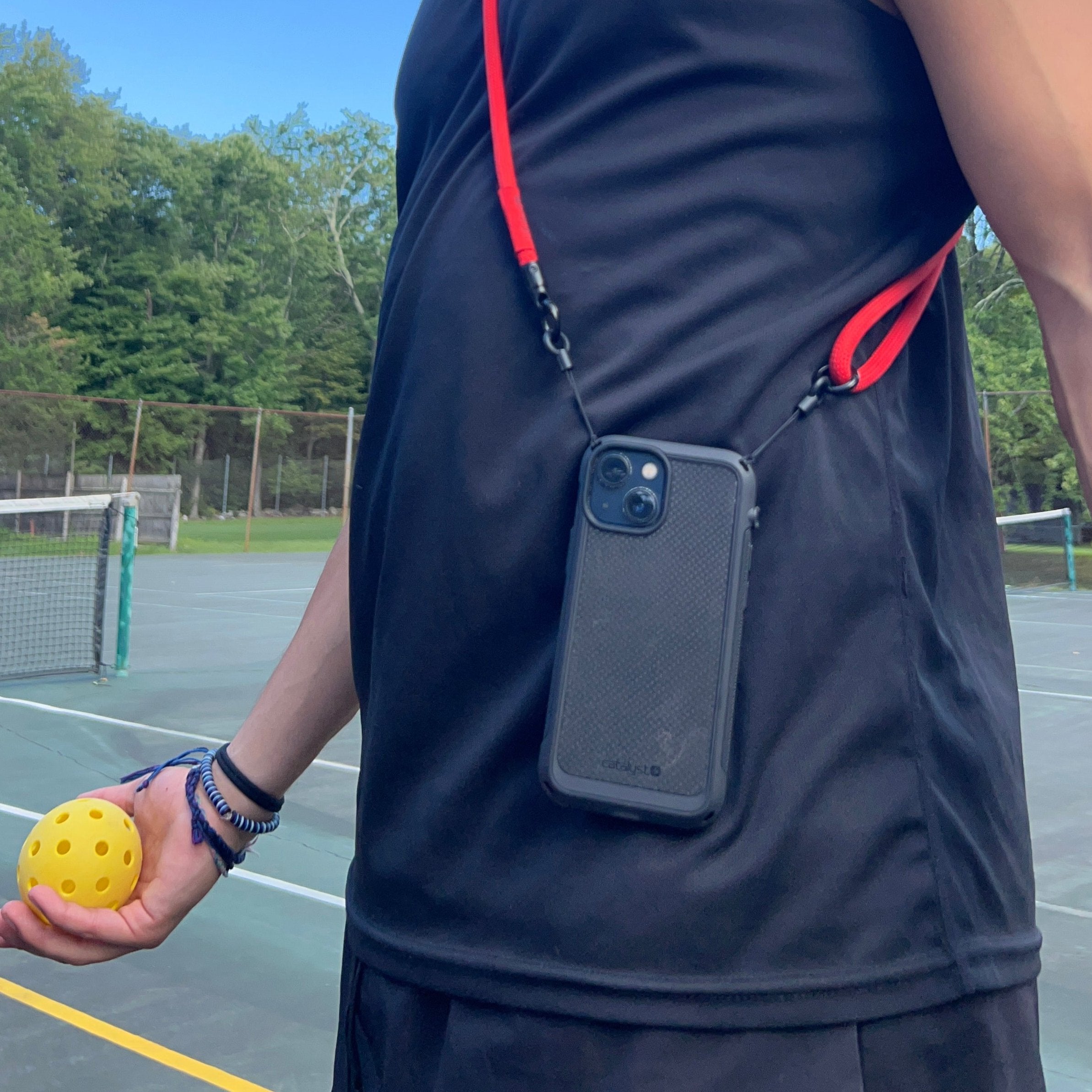 A person is wearing a black tank top and cap while holding a yellow pickleball on a court, with an iPhone 13 in a Catalyst Drop Proof Textured Case attached to their neck by a red strap. Behind them, trees and a blue sky set the scene.