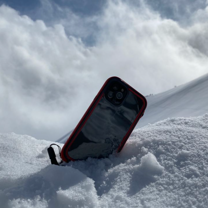 A Catalyst iPhone in a vibrant red, waterproof case offering total protection is partially buried in the snow, tilted against a snowy backdrop under a cloudy sky.