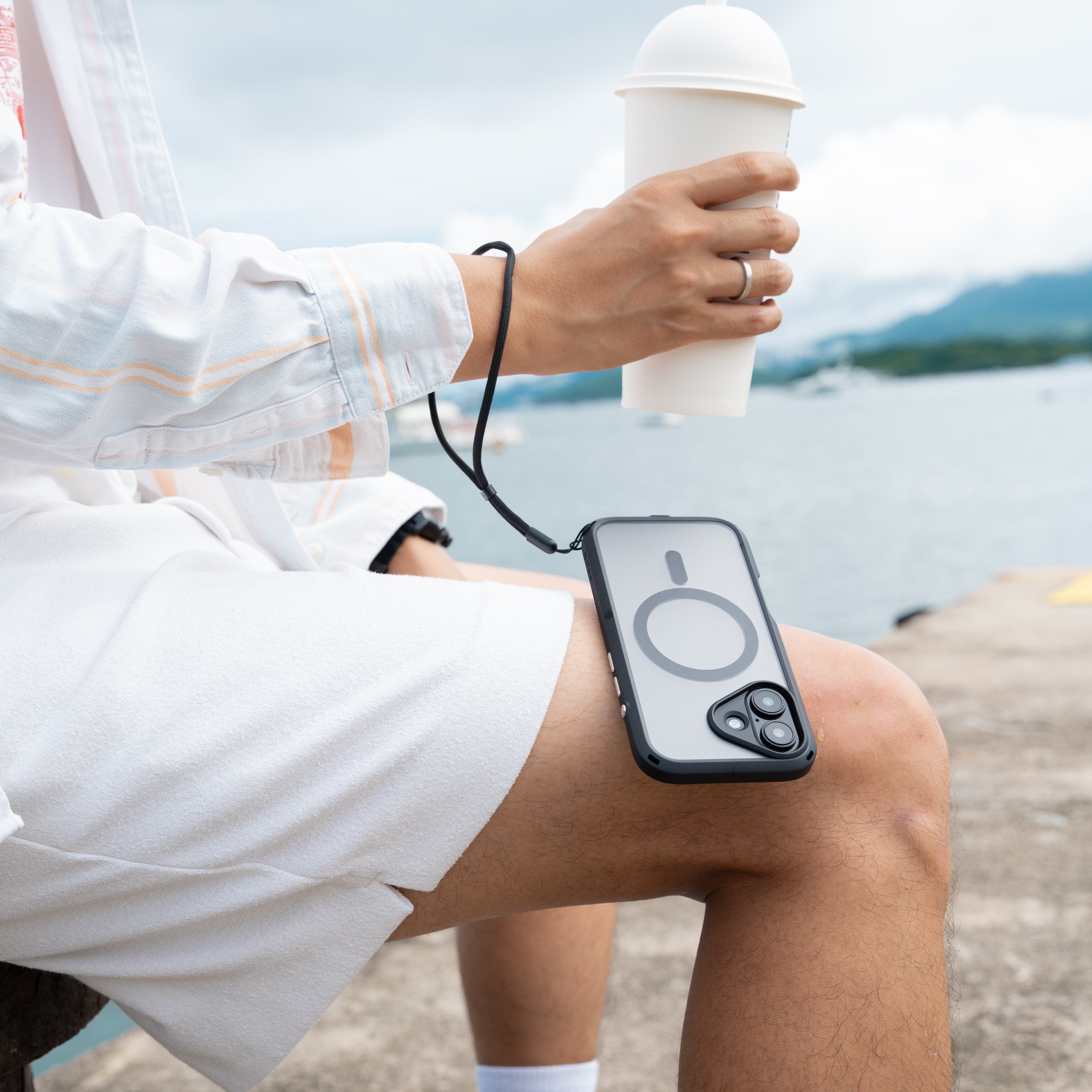 Man seated outdoors, holding a cup with a straw in their right hand and an electronic device in a Catalyst Drop Proof Black Case with a wrist strap resting on their lap. The background shows a body of water and cloudy sky.
