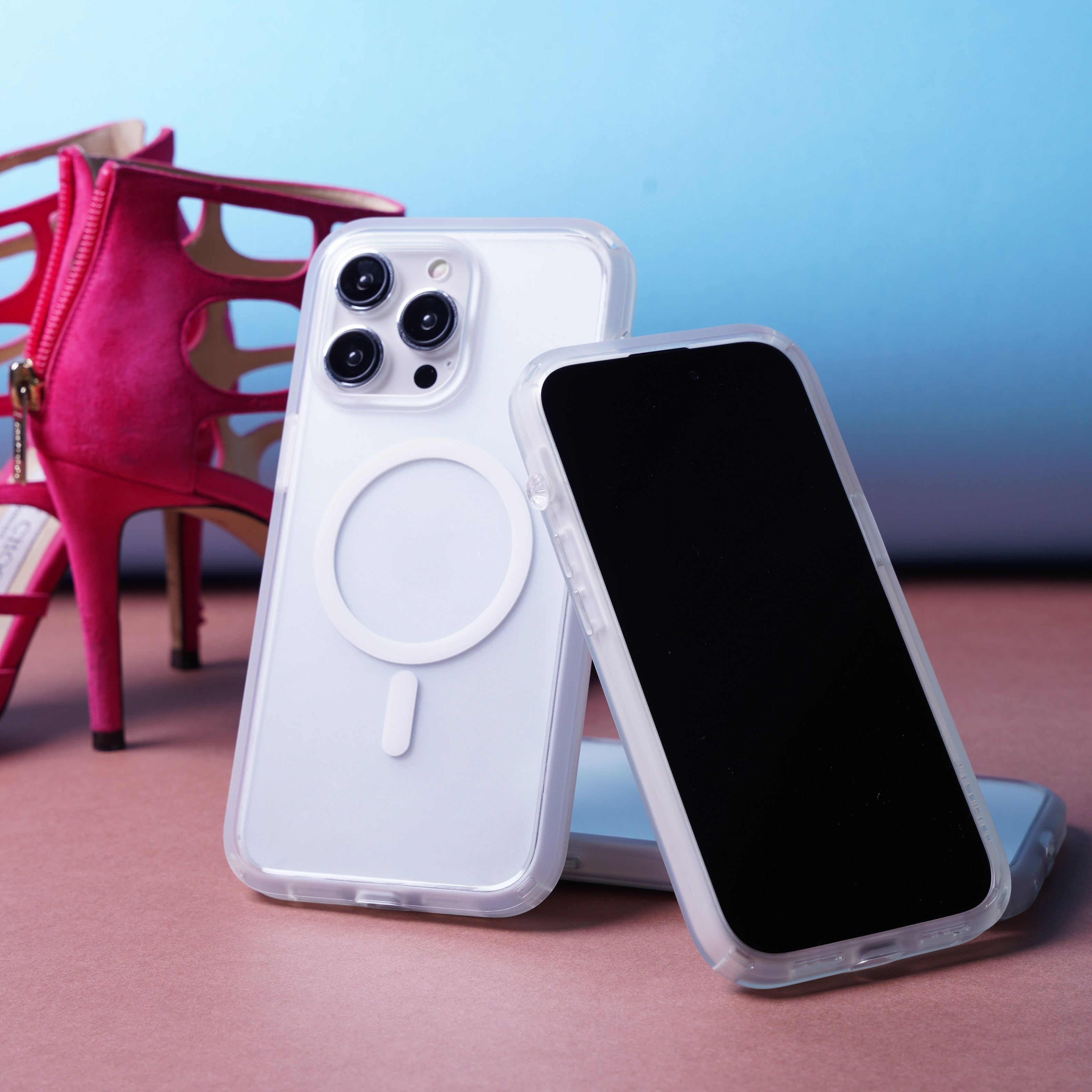 Three smartphones in clear catalyst drop proof cases are displayed on a surface with a high-heeled pink shoe in the background against a gradient blue backdrop.