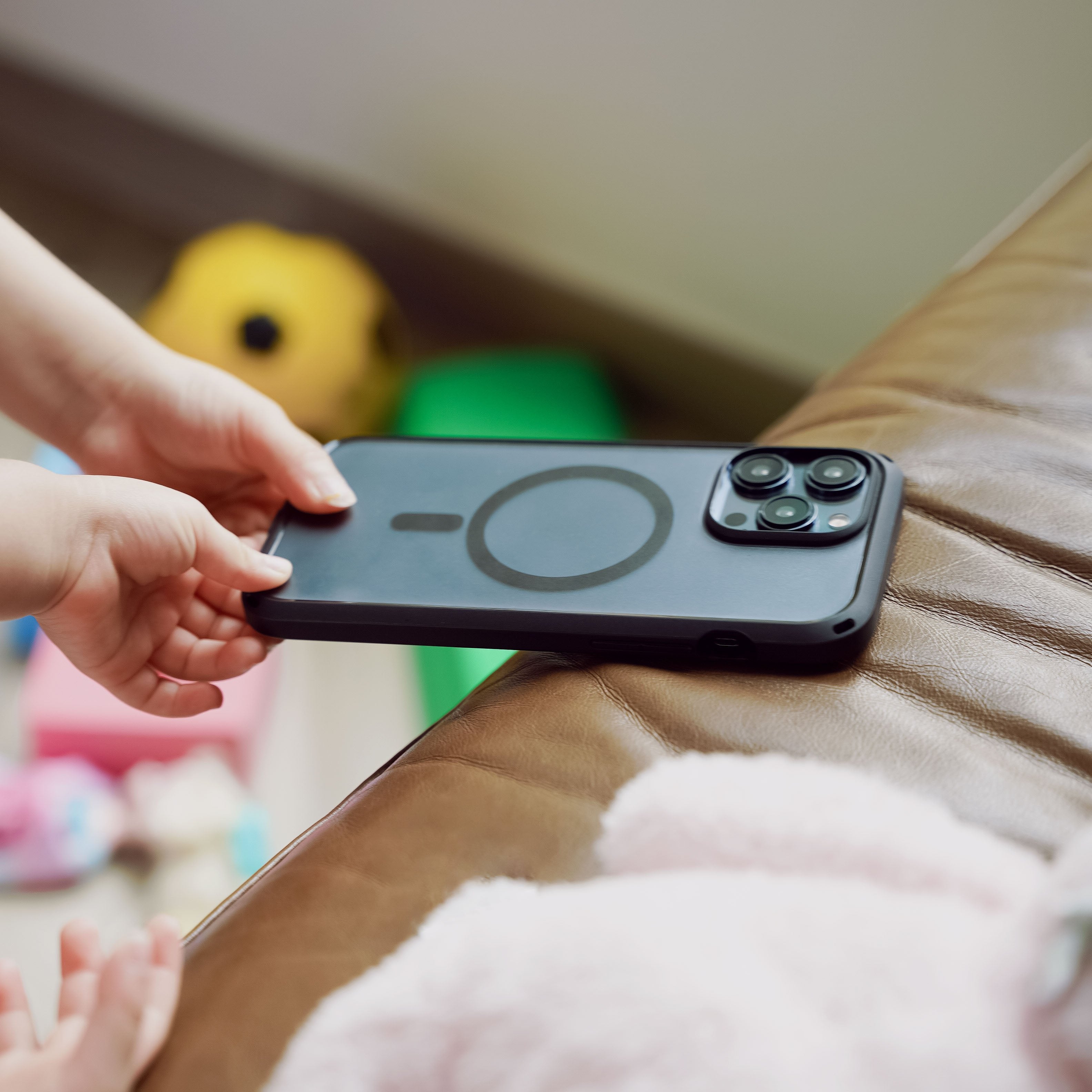 A child's hands reach to pick up a smartphone with a Catalyst Drop Proof Case MagSafe Compatible, resting on a brown leather surface. Colorful toys are scattered in the background.
