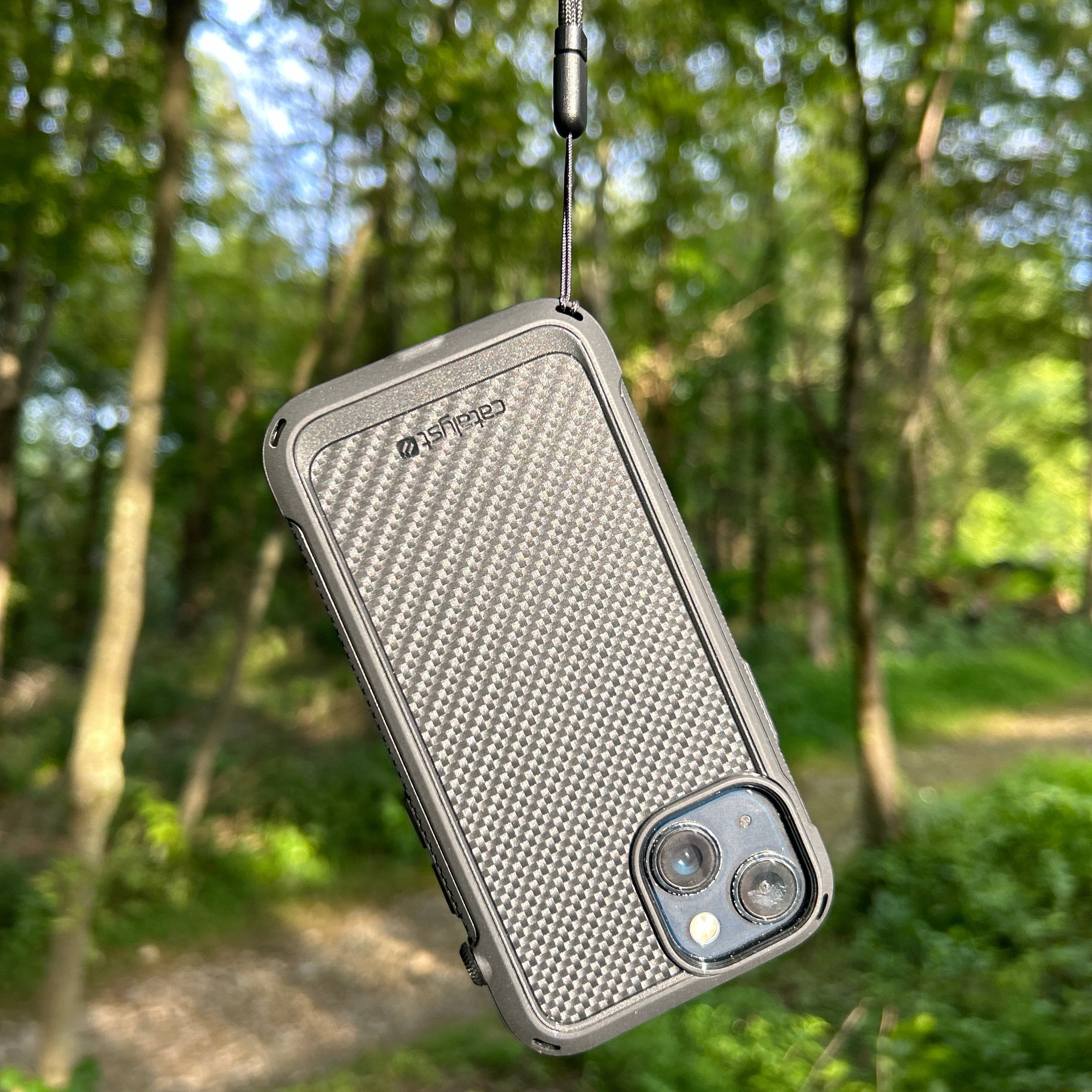 Catalyst iPhone Drop Proof Textured Case in black hangs from a strap in a wooded area, crafted to withstand drops during your adventurous outings.