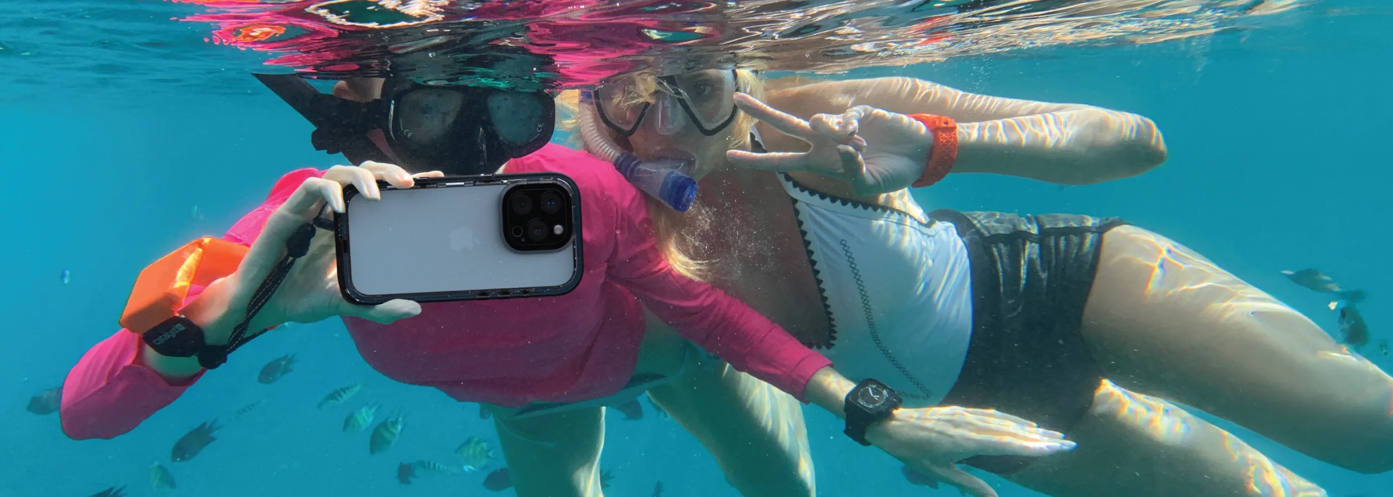 Two snorkelers use an iPhone 16 Pro in a Catalyst black waterproof case, watching as fish dance around them in the clear blue water.